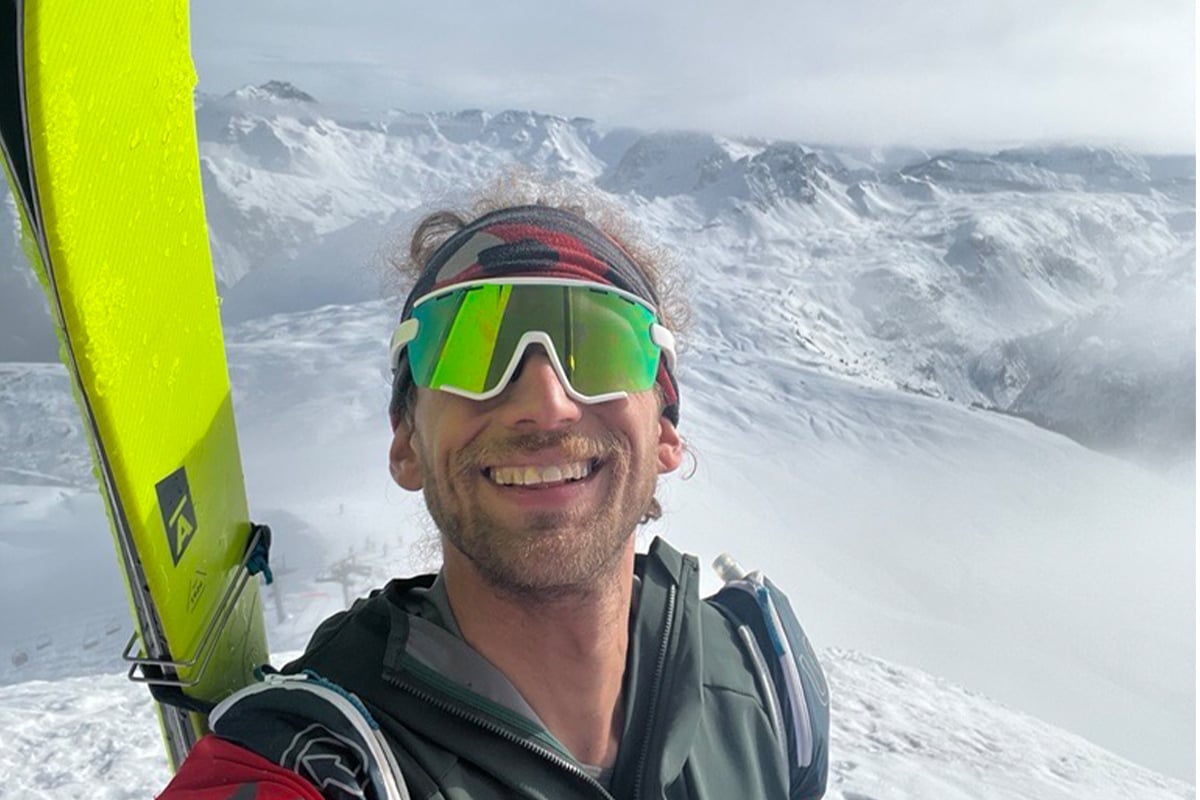Noah Dines smiler på en fjelltopp etter en topptur med ski. Han har på seg solbriller og en Garmin-smartklokke. Snødekte fjell i bakgrunnen.