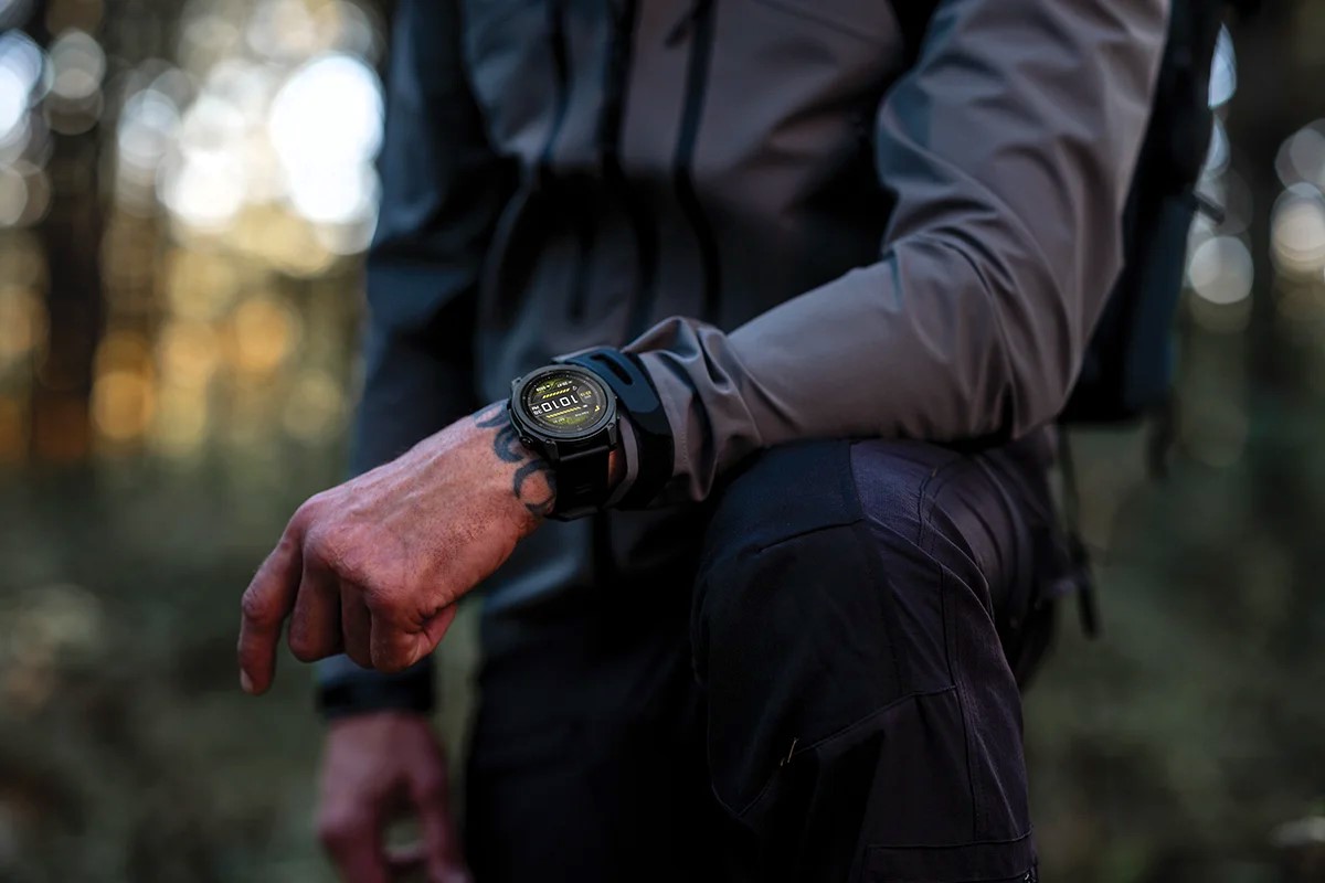 Nærbilde av en person i skogen med en robust taktisk Garmin smartklokke på håndleddet, iført mørke friluftsklær og med armen hvilende mot kneet.