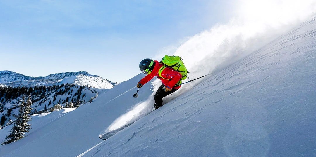 En person kjører på ski ned en bratt, snødekt fjellside i klart vær. Snøen virvler rundt skiløperen, som har på seg rød jakke, svart utstyr og en grønn ryggsekk. Bartrær og fjell sees i bakgrunnen under en blå himmel.